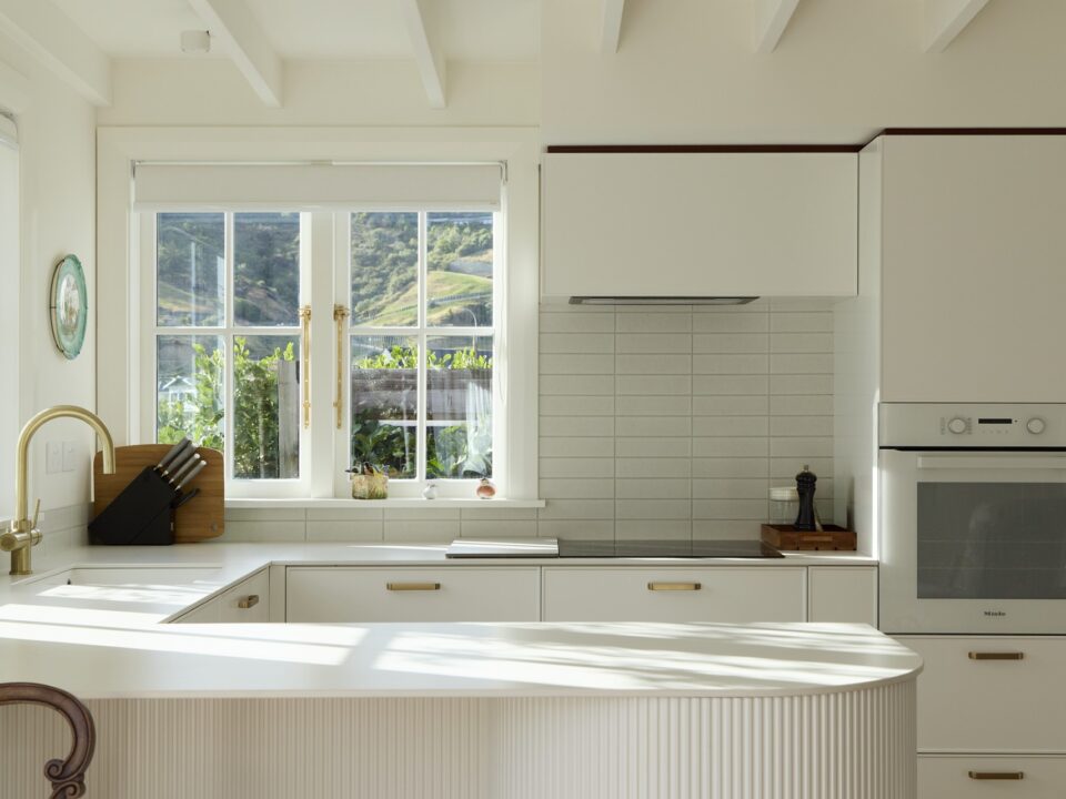 Bright white kitchen with brass tapware, tiled splashback and exposed rafters, lit by afternoon sun.