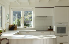 Bright white kitchen with brass tapware, tiled splashback and exposed rafters, lit by afternoon sun.