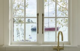 Brass mixer tap and white sink beneath a timber-framed sash window looking out to the estuary and moored boats.